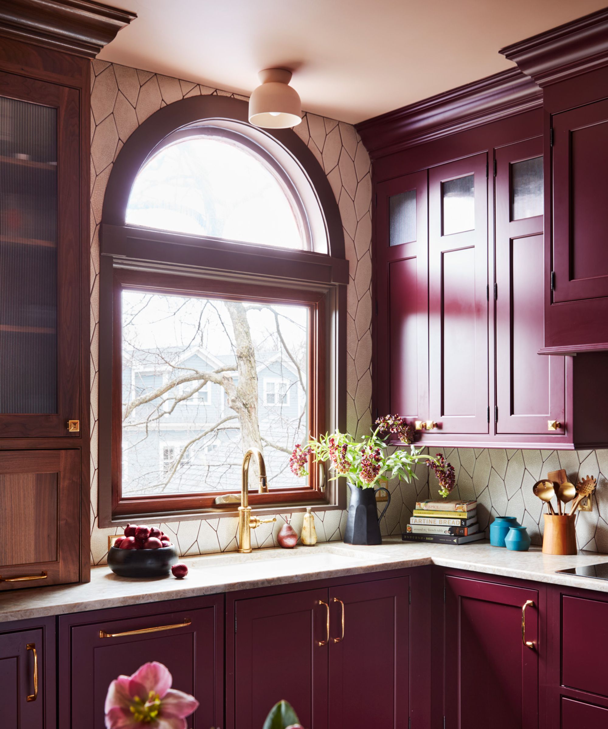 The corner of a kitchen with burgundy cabinets, a plaster pink ceiling, white countertops, and an arched window.