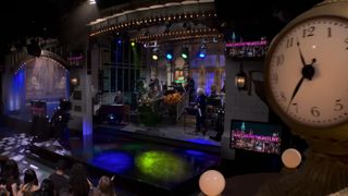 The opening shot of the Saturday Night Live studio with the iconic clock in the foreground and the stages behind it with the band playing and a crowd waiting for the host. 