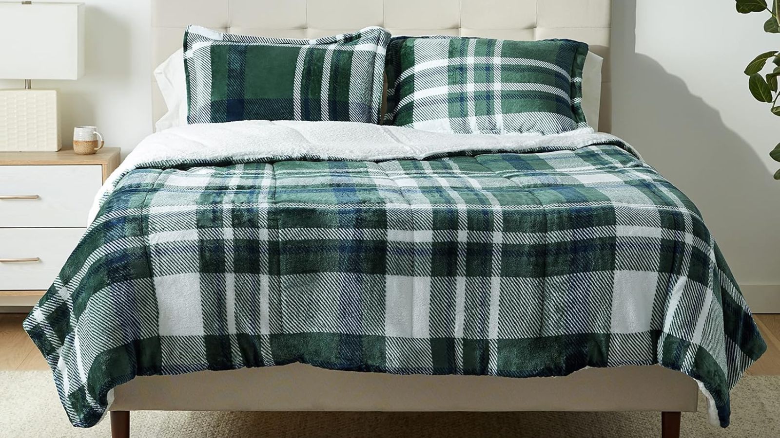 A green and white plaid bedding set on a cream double bed, in a neutral bedroom with a cream, and wood bedside table, a white lamp, white walls, and a large plant to the right.