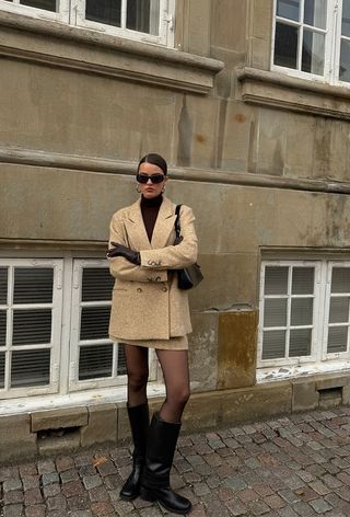 The winter capsule wardrobe item, a wool blazer, is shown in a photo of a woman standing in the street wearing a brown turtleneck under a tan blazer with a matching mini skirt, black tights, leather gloves, and black knee-high boots