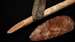 A black projectile point, a reddish stone blade and a brownish rod against a black background.