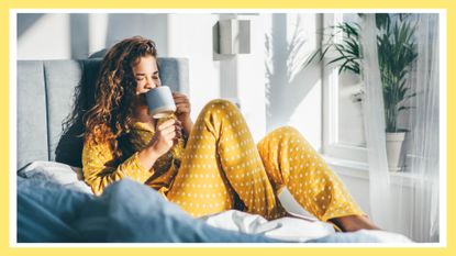 Woman in yellow pajama sitting on bed and drinking coffee at the sunny morning bedroom, healthy morning routine