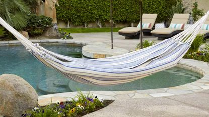 A blue and white striped hammock hanging in a paved backyard by a swimming pool