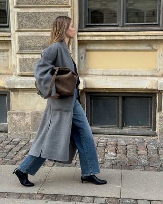 Danish art director Clara Dyrhauge poses on a cobblestone sidewalk wearing an oversize gray belted coat, a brown suede bag, straight-leg jeans, and black kitten-heel boots