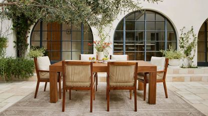 best outdoor furniture. Outdoor courtyard, wooden dining table with six seats, stone flooring, greenery hanging from house in background, striking arch windows