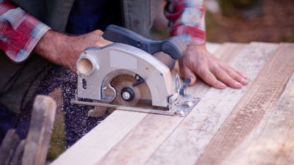 Close up of man in plaid red and blue long sleeve shirt using a circular saw on a smooth piece of long wood