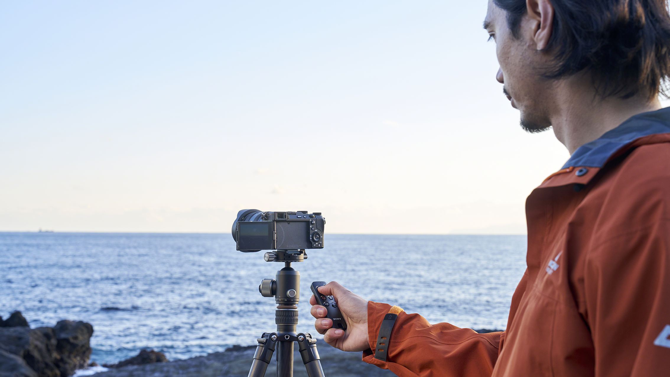 A person wearing an orange jacket operates a camera on a tripod with Sony RMT-VP2 remote control, aiming at the ocean under a clear blue sky