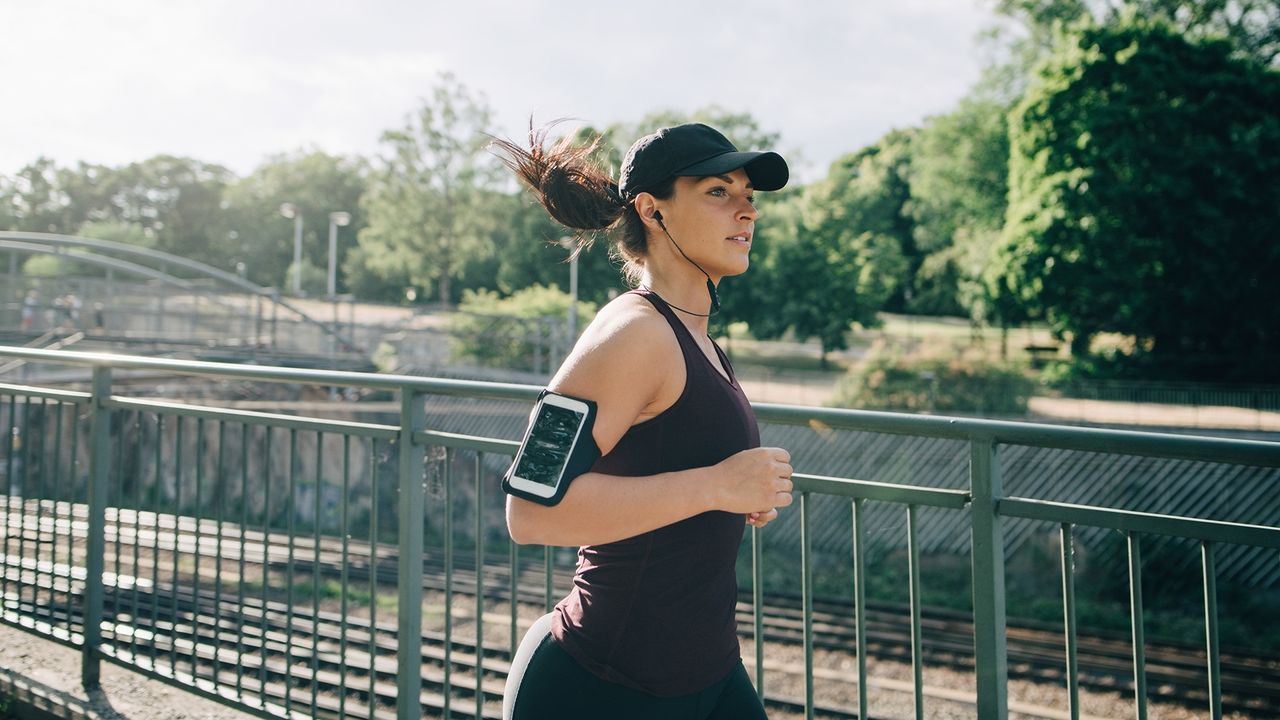 Confident sportswoman listening music through in-ear headphones while jogging on bridge in city