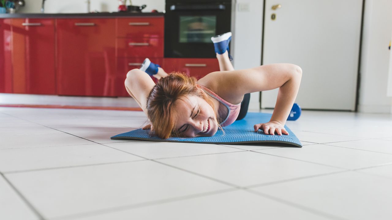 Woman smiles as she lies face down on an exercise mat on a tiled kitchen floor