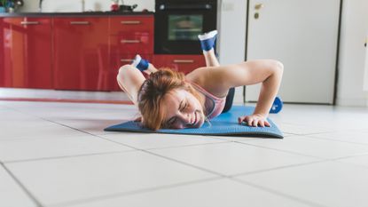 Woman smiles as she lies face down on an exercise mat on a tiled kitchen floor