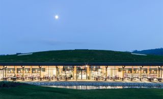 Night time image, building set into a hill, grass verge to the front with a rocky pond, glass fronted, lighting, a few people sat at the table and chairs outside, blurred furniture inside, grass roof with skylights, trees in the backdrop, clear night sky with blurred moon in the distance