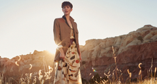 A woman stood in long grass wearing clothing available from Banana Republic with the setting sun and a rock formation behind her.