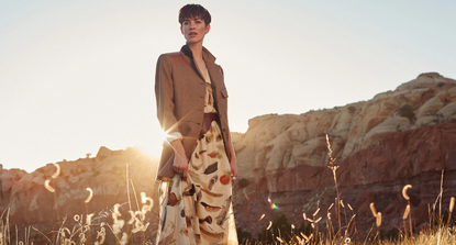 A woman stood in long grass wearing clothing available from Banana Republic with the setting sun and a rock formation behind her.