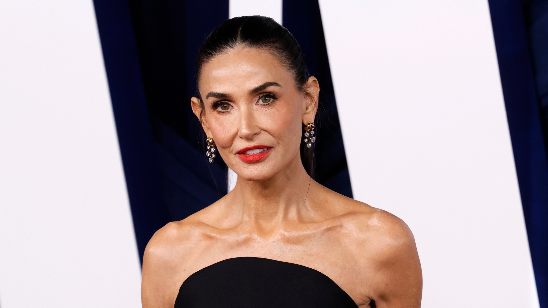 Image of Demi Moore with her black straight hair tied into a sleek ponytail and wearing a black strapless top, crystal drop earrings and red lipstick