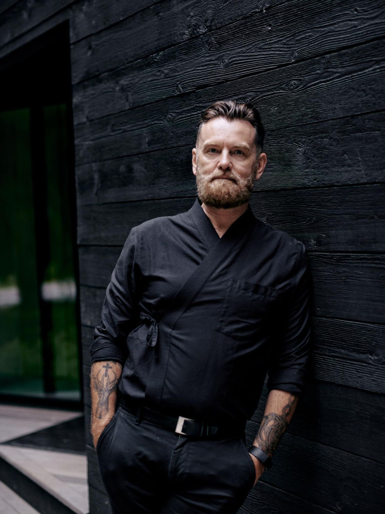 A man dressed in a total black outfit with his hair pushed back stands with his hands in his pockets against the wooden outdoors of a house.