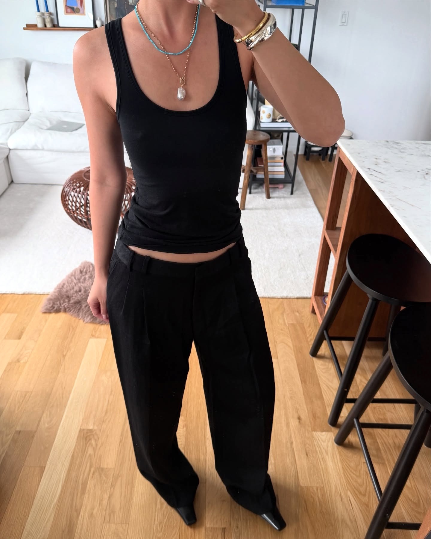 Style influencer Mimi Nguyen poses for a mirror selfie in her NYC apartment wearing a turquoise beaded necklace, a pendant necklace, a black scoop-neck tank top, black pants, and square-toed shoes.