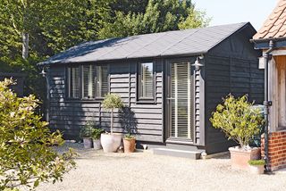 a large garden barn painted black on the outside with white shutter blinds, and potted plants on the outside