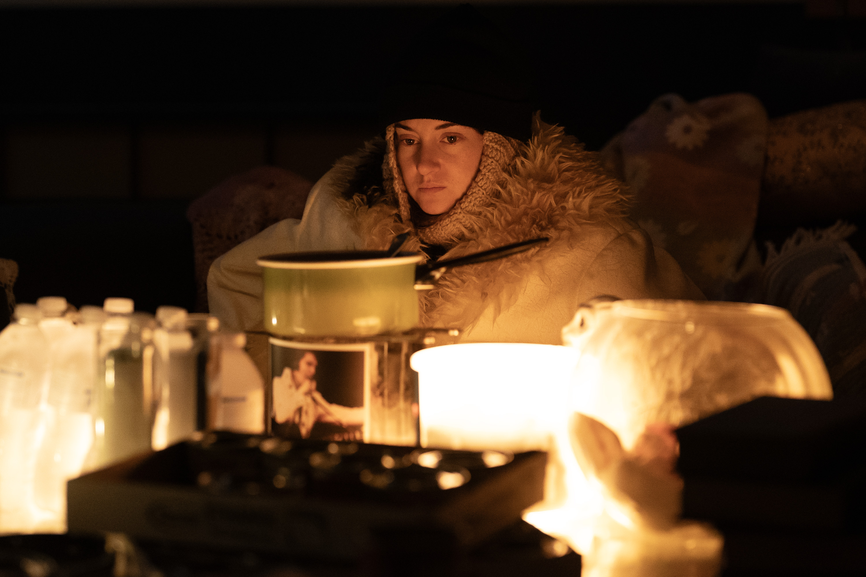 A woman (Shailene Woodley as Annie Clay) wears a black beanie and a fur-lined blanket as she sits in a dark room lit with candles. A burner adorned with an Elvis photo and a pack of water bottles is seen on a table. A still from 'Paradise' season 2.