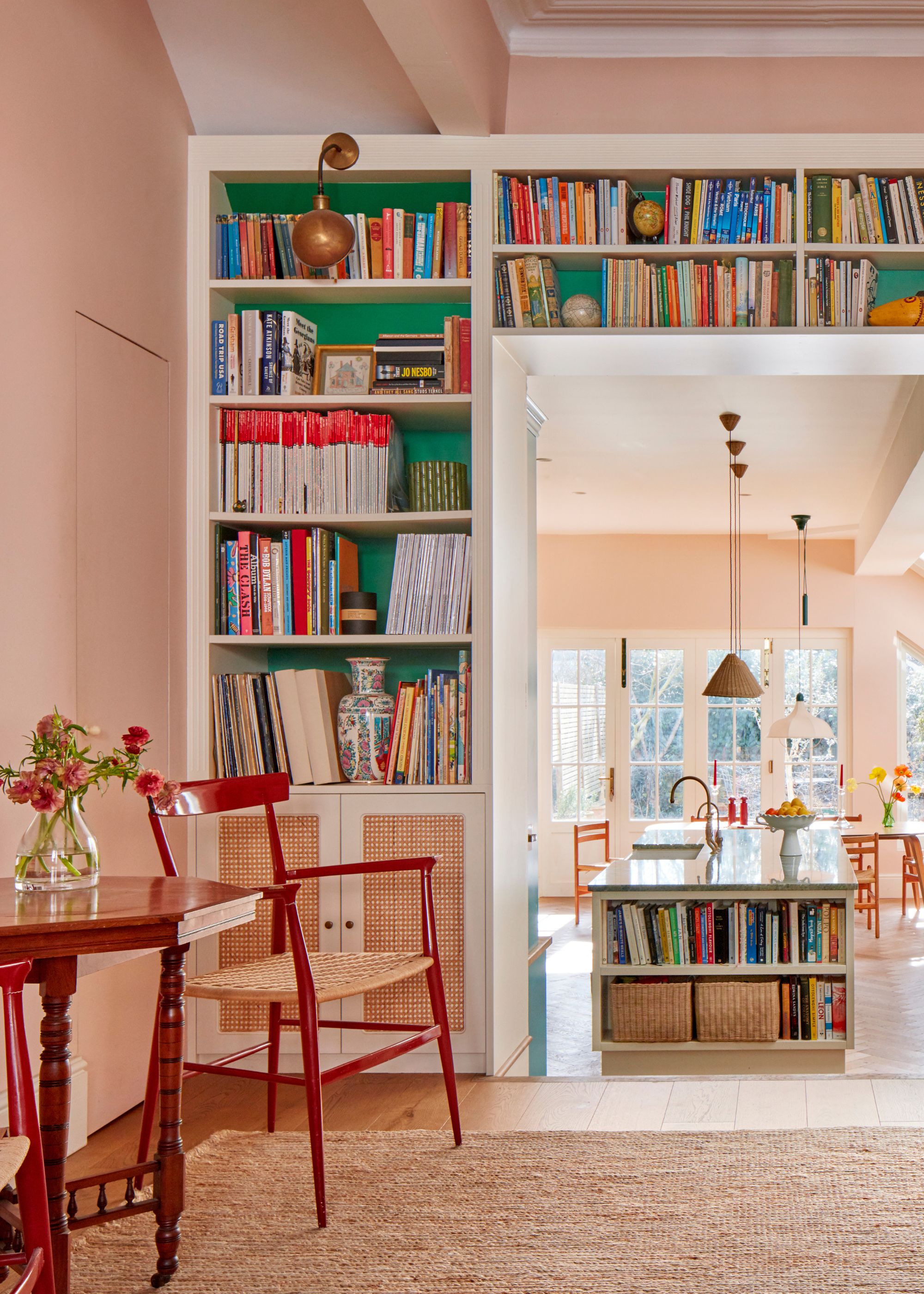 A living room with pale pink walls, large bookshelves around the room with bright green on the backs, a jute rug, and a side table with two red chairs.