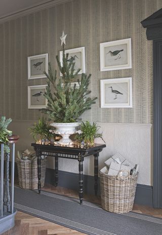 A hallway decorated for Christmas, with garlands made of greenery on the staircase, a console table and small Christmas tree and wicker baskets full of presents