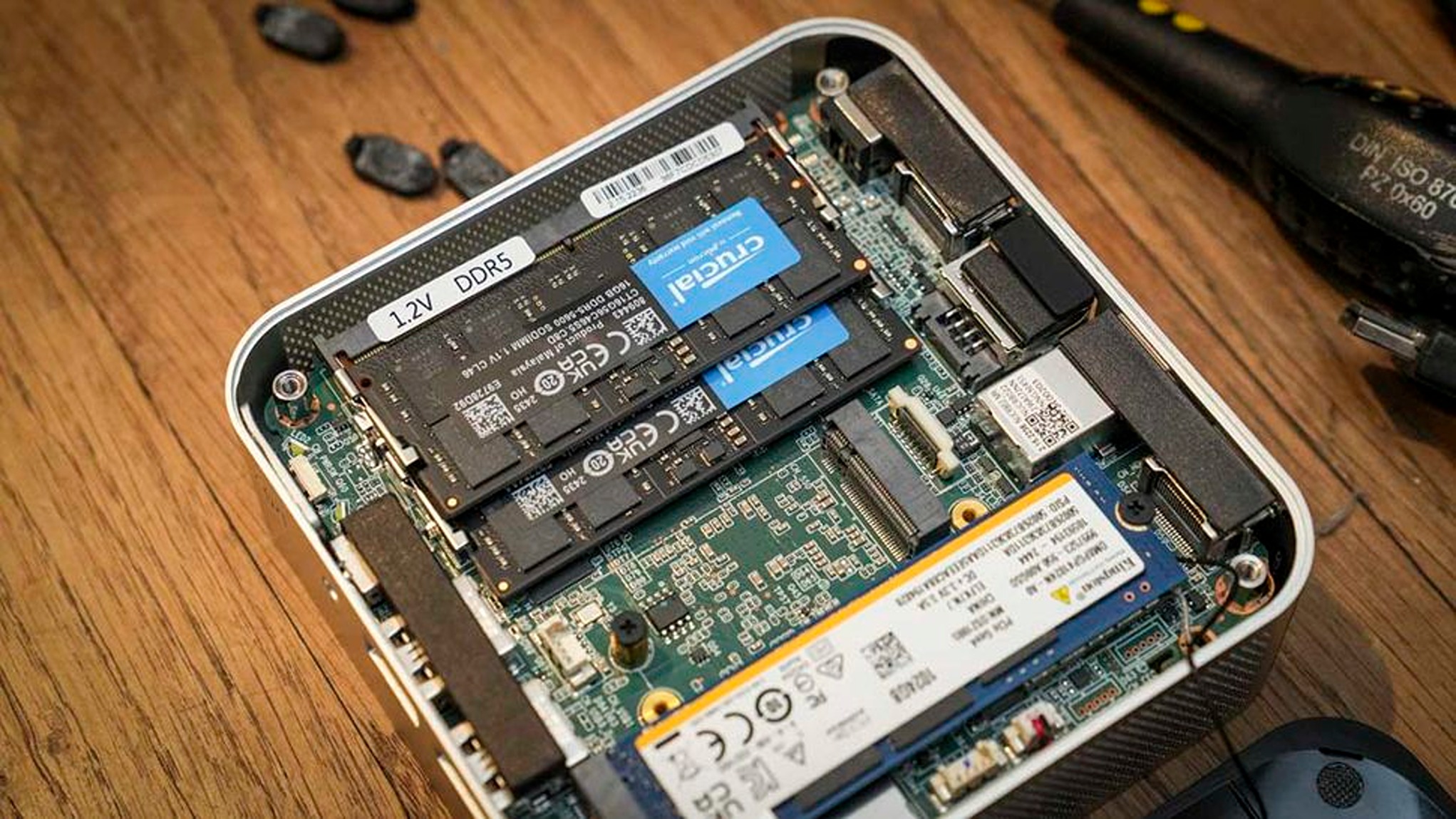 Open computer case on wooden table showing exposed circuit board, RAM labeled "12V DDR5," storage components, and screws nearby; a small screwdriver is partially visible, conveying a DIY upgrade scene