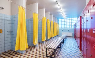 Changing room featuring individual shower cubicles with yellow shower curtains, red lockers opposite, a line of benches and hairdryers on the end wall