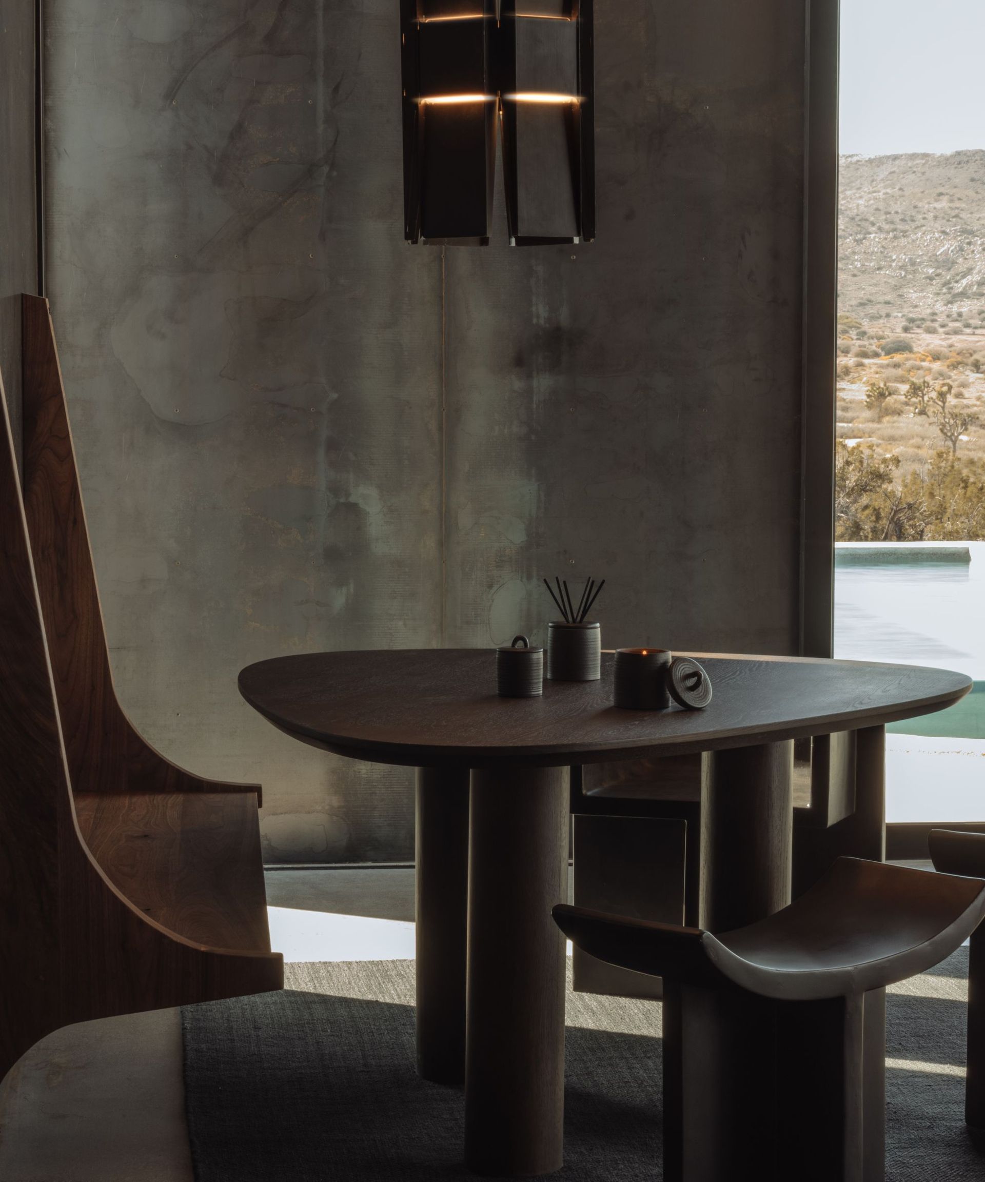 An Adlan Fragrances diffuser on a wooden table with wooden chairs, against an exposed, industrial style wall. To the right is a window with a view of a swimming pool and valley.