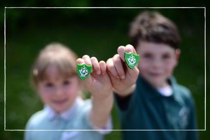 two school age children, one boy and one girl, in school uniforms holding green Blue Peter badges towards the camera