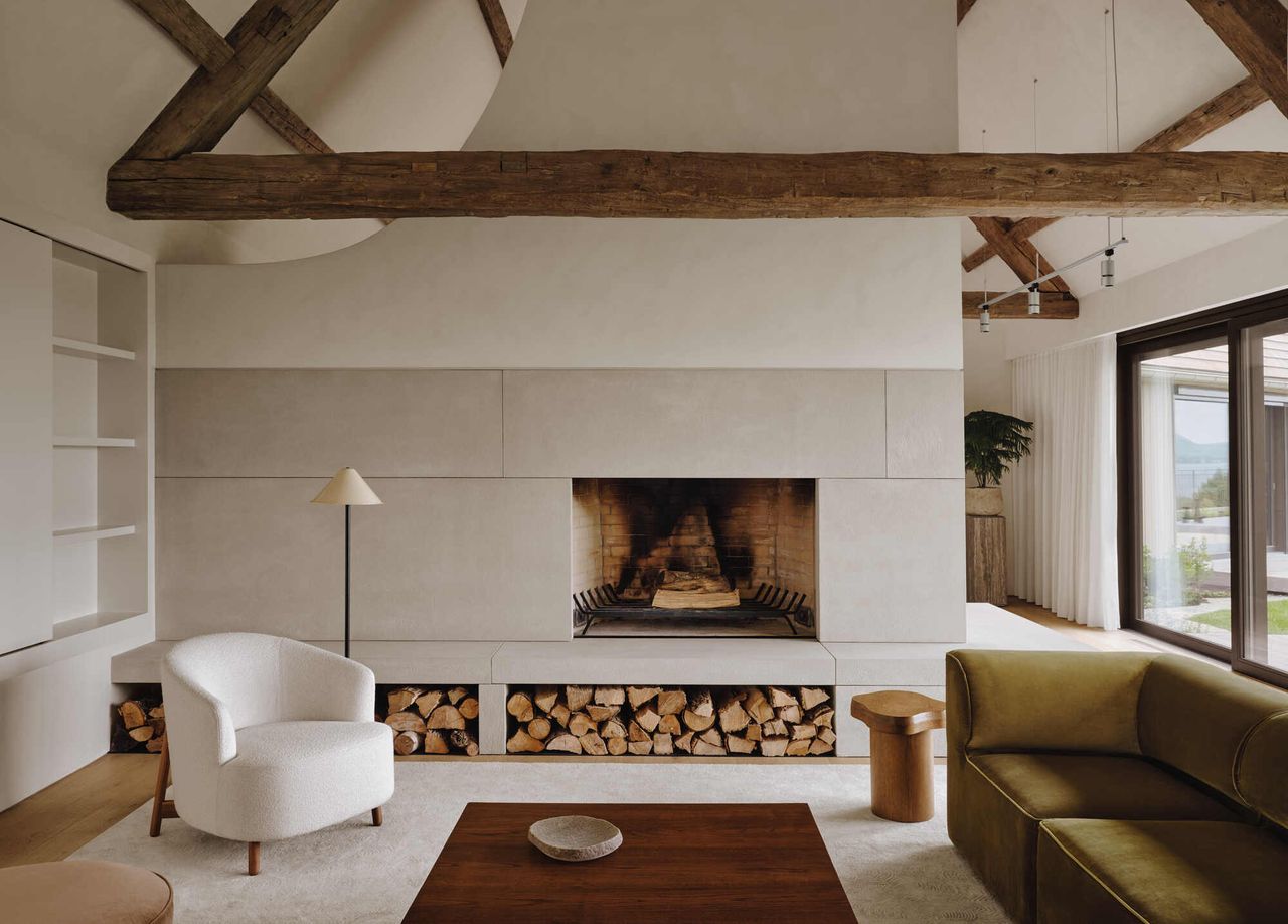 modern living room with built-in fireplace with logs stacked, a white armchair, green sofa, and exposed timber ceiling beams