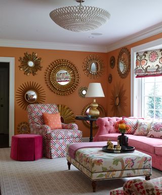 living room with bright orange walls, pink sofa, patterned armchair, gallery wall with maximalist gold mirrors