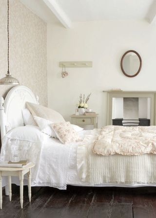 white bedroom with pale pink wallpaper, floral quilt, peg rail, painted side table and matching fire surround, lace pillowcases, painted white bed, vintage pieces