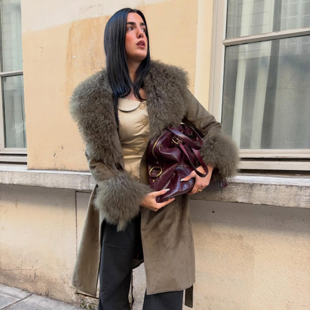 An image of an influencer in Paris wearing an olive green fur coat with a button-down top and holding a burgundy leather tote