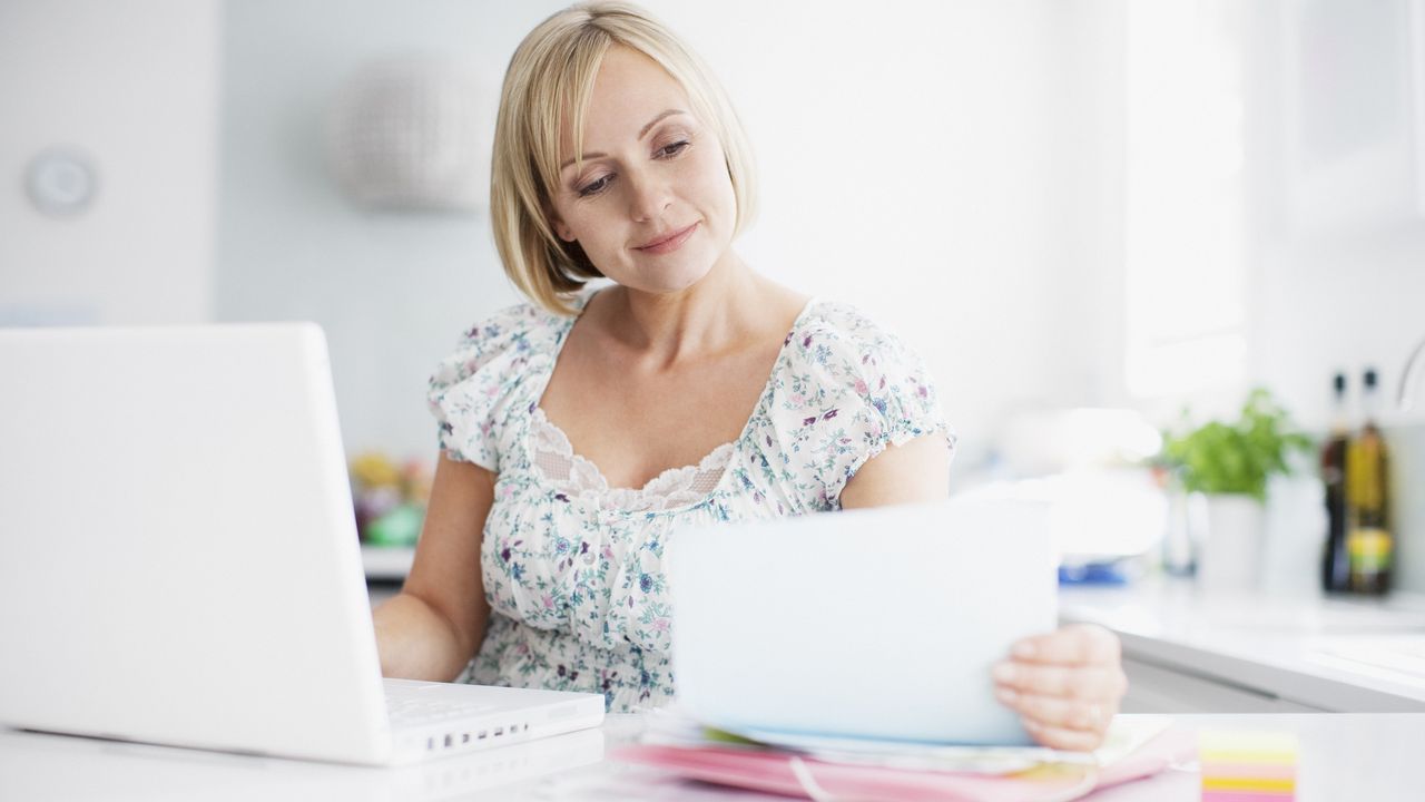 A woman works on financial paperwork while sitting in her kitchen at her laptop.