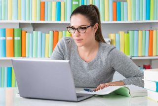 Female teacher looks at laptop computer with open book and pen on table.