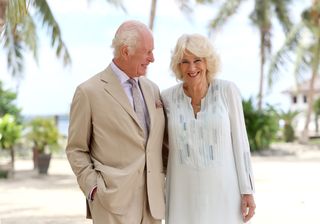 King Charles wears a tan suit while smiling at wife Queen Camilla, who wears a light blue tunic with a striped pattern, while they pose on the beach in Apia, Samoa