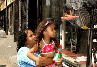 A lady holds a child up to a fan providing some relief on a hot day.