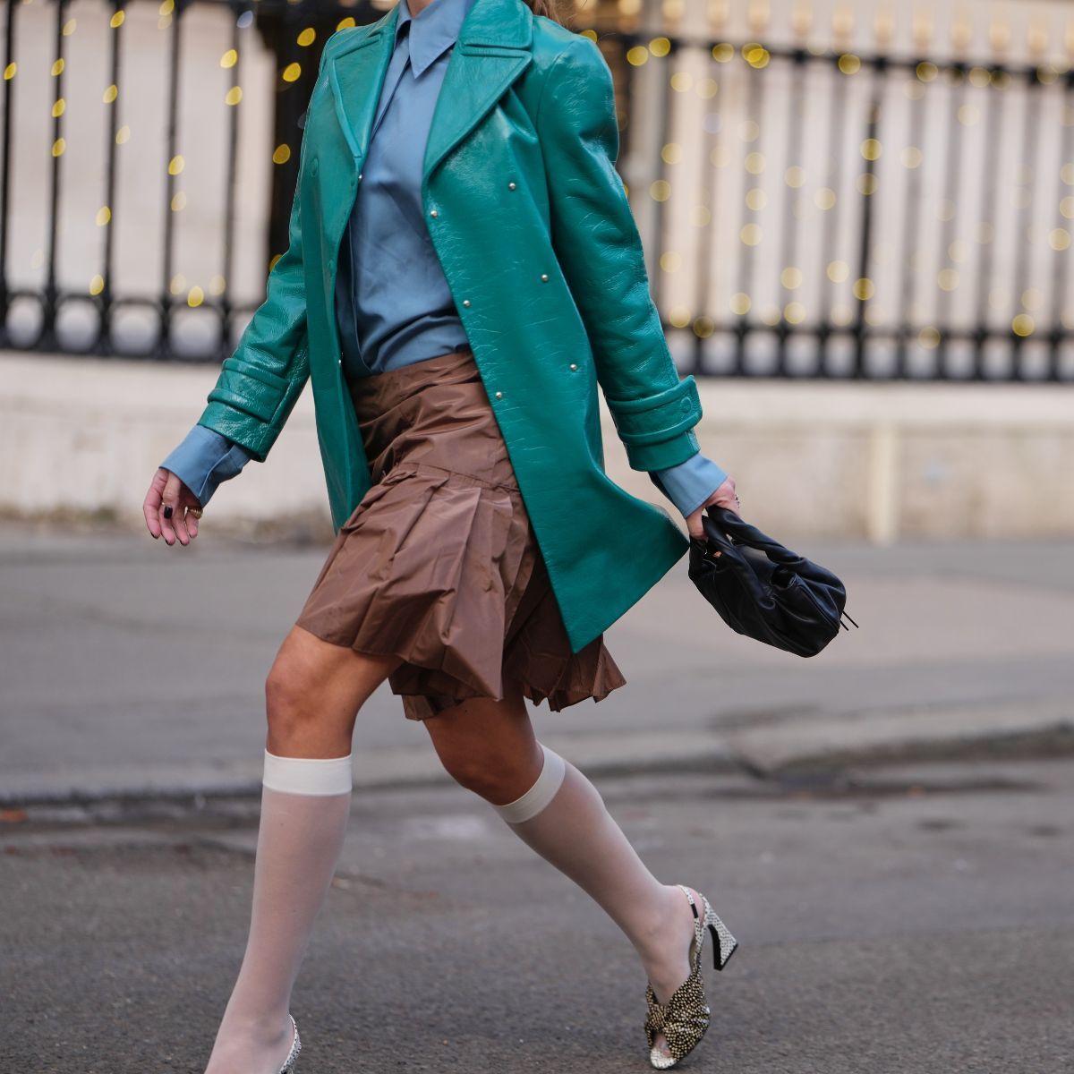 street style shot of woman wearing turquoise trench, blue shirt, brown nylon skirt and white sheer knee high socks