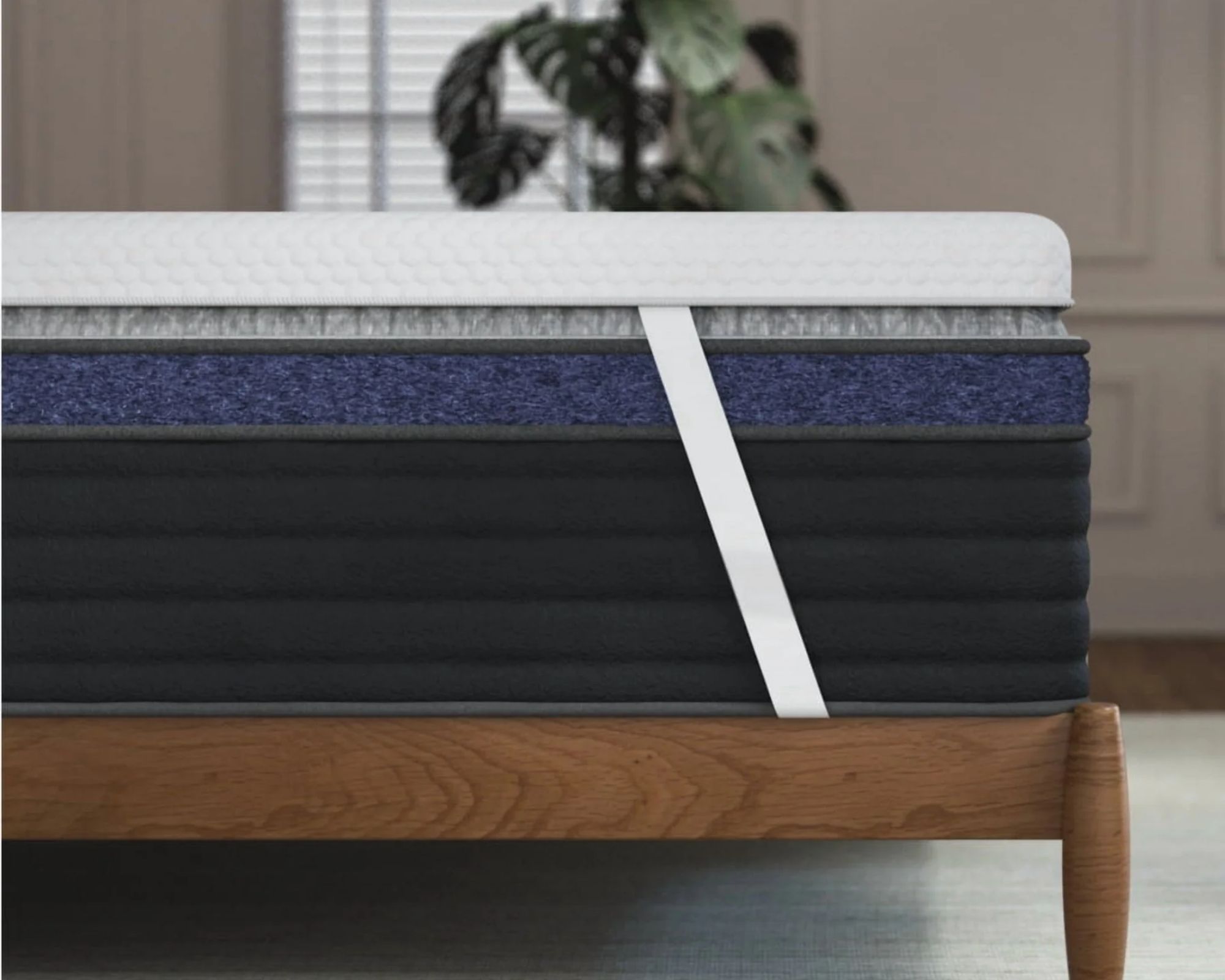 The white Helix ErgoAlign Mattress Topper with white corner straps is seen on a the corner of a navy mattress on top of a wooden bedframe. The floor of the room is grey, and behind is a neutral panelled wall, large cheeseplant and window with blinds. Everything beyond the bed is out of gocus.