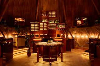 A warmly lit cocktail bar inspired by music culture is housed within a chapel-style wooden building, with sleek finishes, plants, and a back-lit cocktail display.