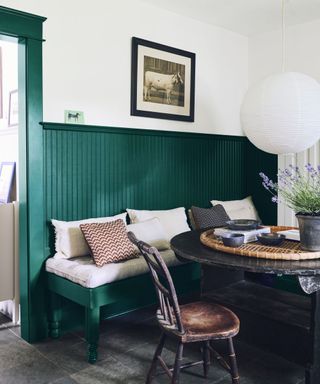 A dining room with a white wall with a wall art print with forest green wall panels below it with a white cushioned bench, a black circular table with a rattan board and a brown wooden chair next to it