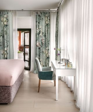 A bedroom with a dressing table, pink bed and printed floral curtains
