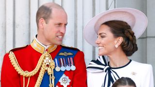 Prince William (wearing his Welsh Guards uniform) and Catherine, Princess of Wales (wearing a white hat and dress) look at each other on the Buckingham Palace balcony at Trooping the Colour 2024