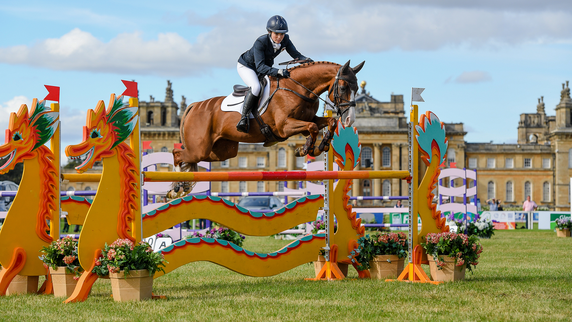 Georgia Bartlett, a rider in a blue coat, rides chestnut horse Spano De Nazca over an orange showjump with wavy planks, in front of Blenheim Palace