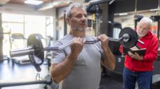 A man curls an EZ-curl barbell up to his chest in a gym, while a trainer watches him and makes notes in a book. 