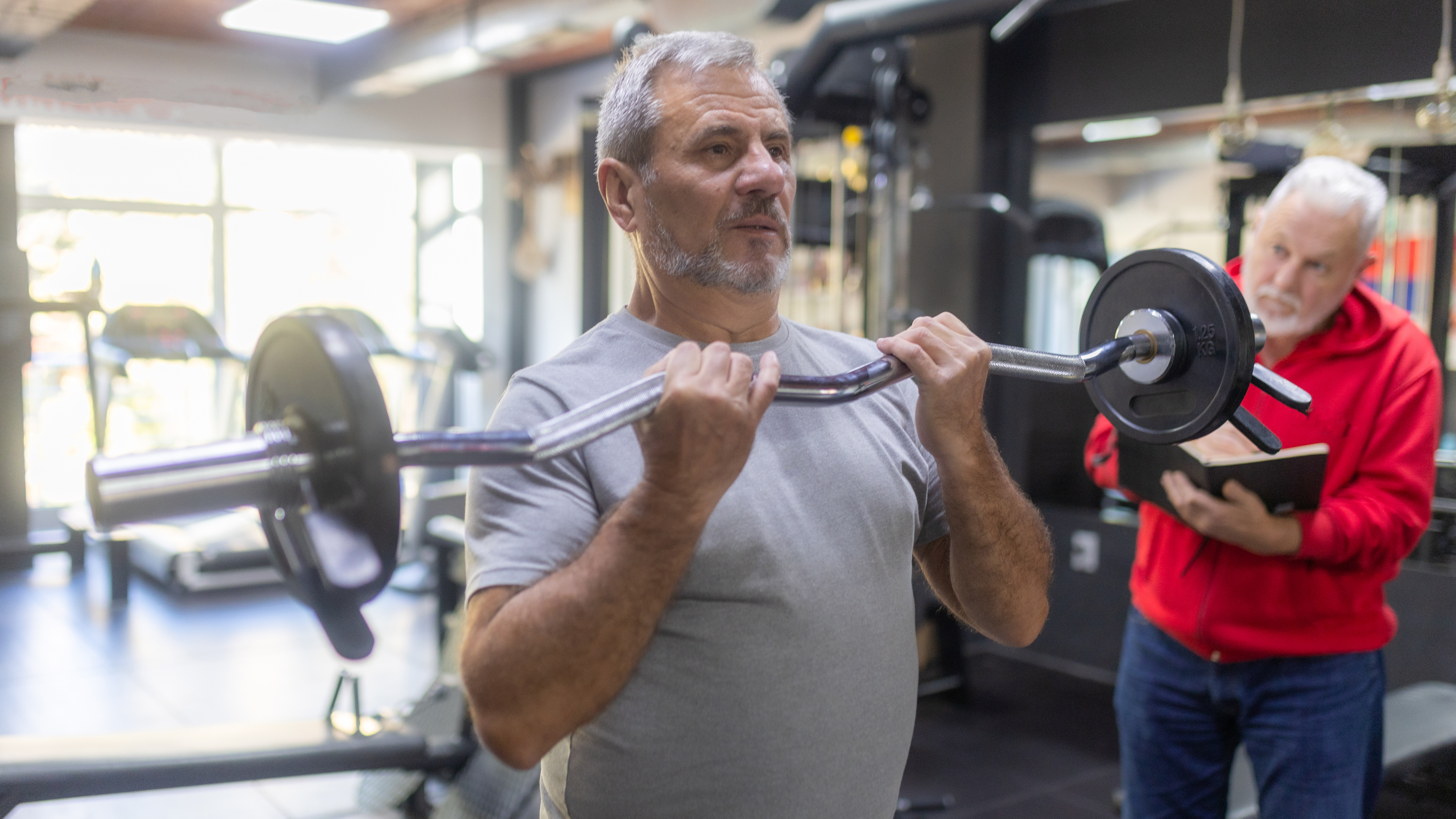 A man curls an EZ-curl barbell up to his chest in a gym, while a trainer watches him and makes notes in a book. 