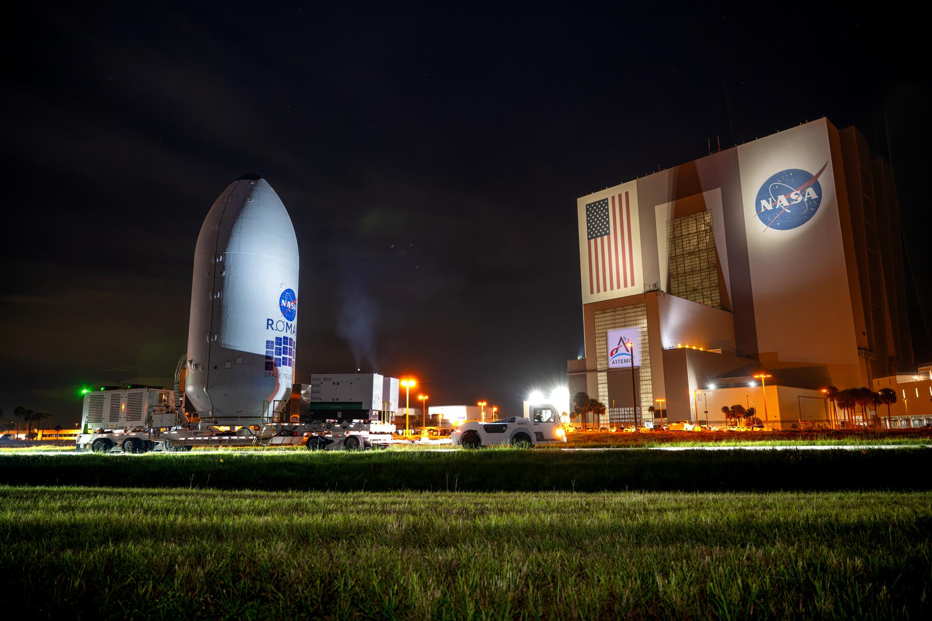 A white, rounded point cylyictrical capsule is rolled by a giant cube building.