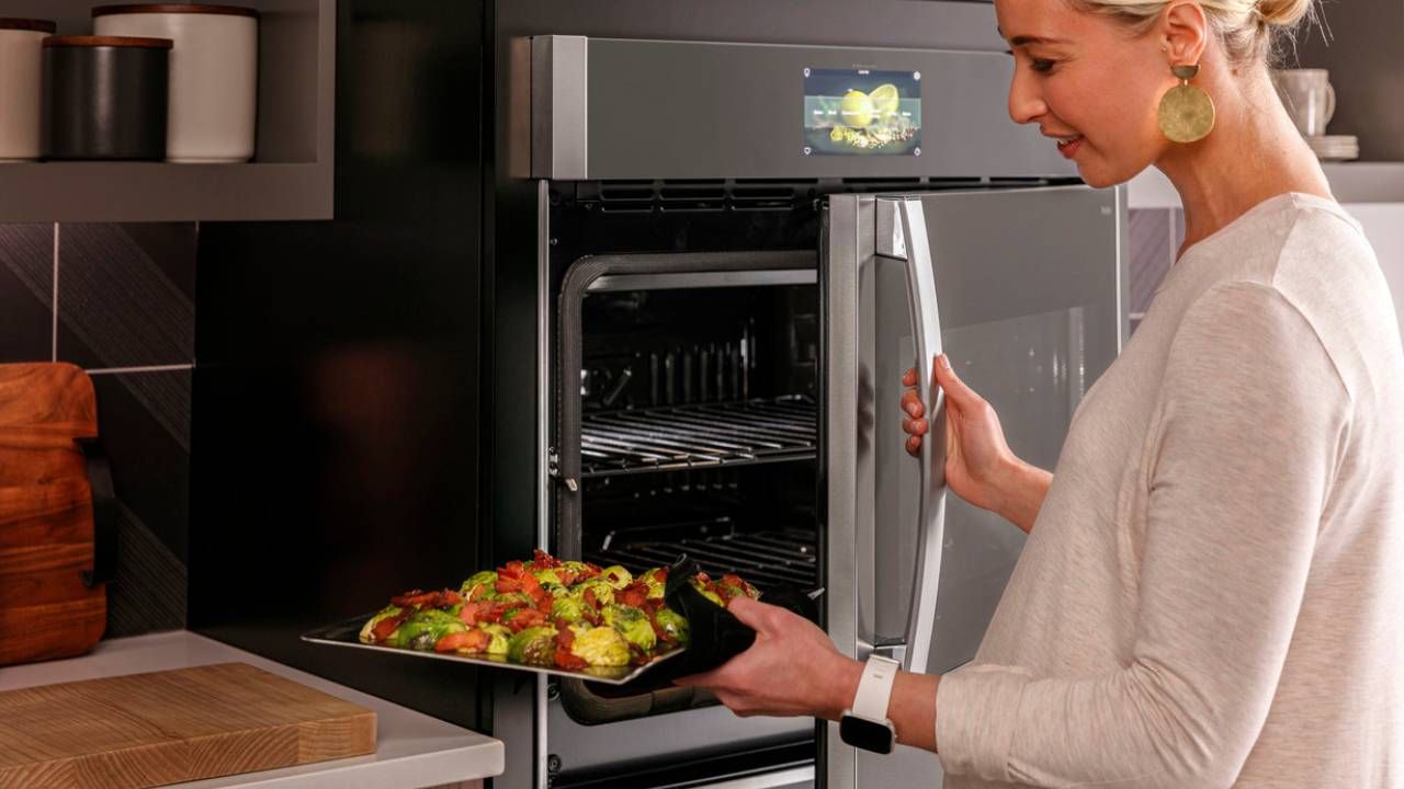 A woman holding a tray of vegetables and opening the door of a GE Appliances wall oven