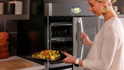 A woman holding a tray of vegetables and opening the door of a GE Appliances wall oven
