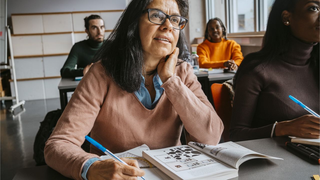 An older woman sits in a classroom with a textbook open in front of her.