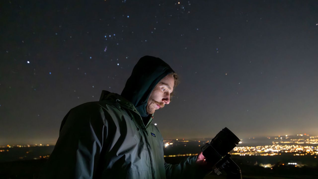 A man looking at the Fujifilm X-M5 at night with Orion's Belt and city lights in the background